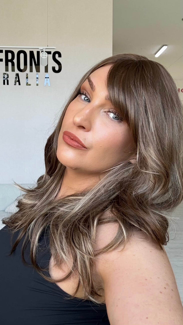 20 inch warm brunette hair with ash and honey blonde highlights, a long fringe parted to the side and light layers throughout.