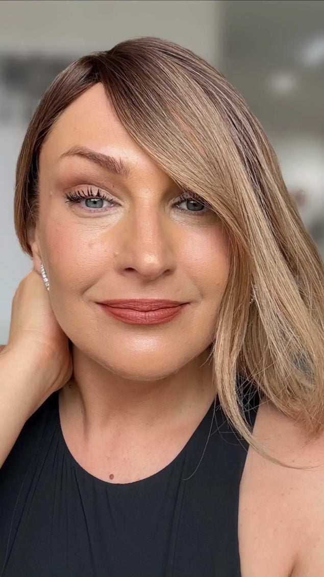 a close up shot of natural looking hairline with face framing layers slightly over one eye, slight side part, with soft brunette root and light curl throughout.