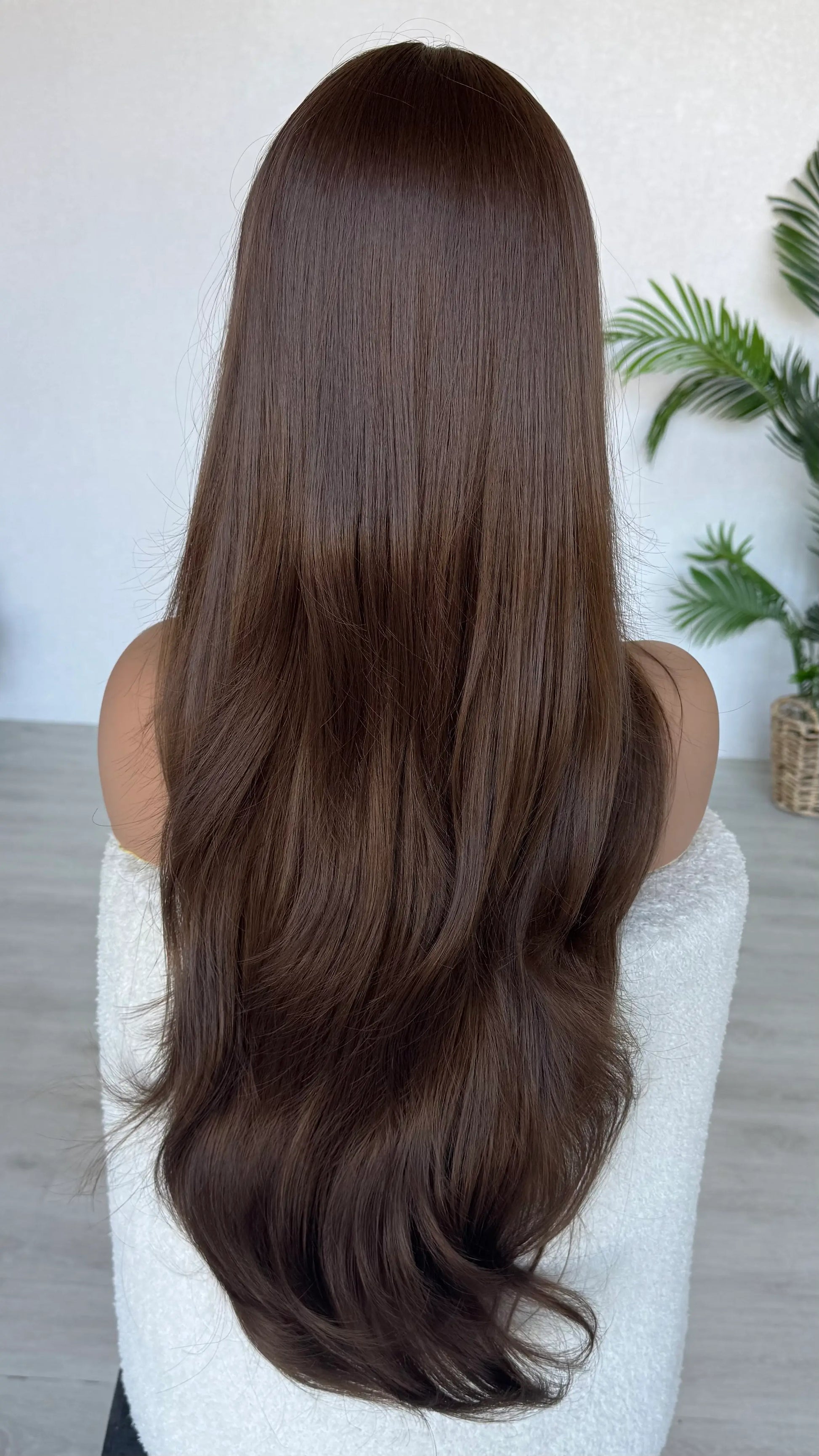 Person with long, straight brown hair sitting on a white chair indoors. australia wigs.