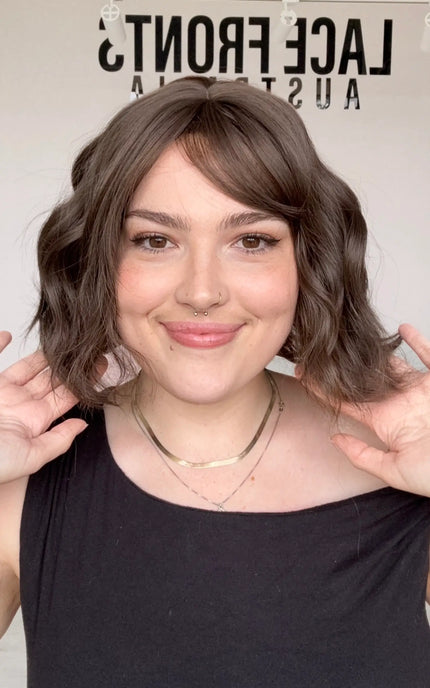 a front shot of the wavy neutral to cool toned brunette hair, wavy styling with the middle part and fringe parted to the right hand side, perfect for daily wear with a glueless cap and heat safe fibres.