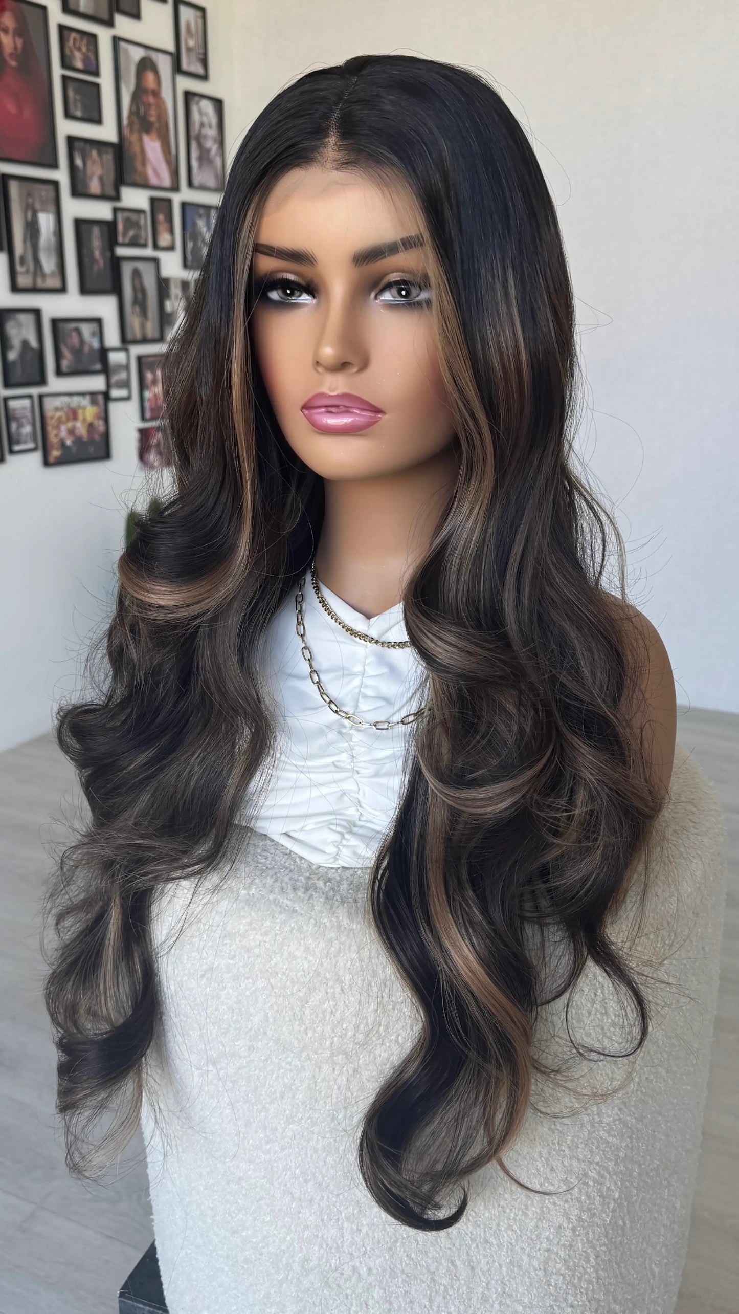 Mannequin head with long, wavy brown wig against a wall with photo frames. brunette hair with highlights, wavy hair.