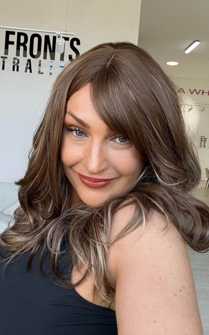 Woman smiling in a warm brunette wig with honey blonde highlights, worn with a middle part and a long fringe parted to the side.