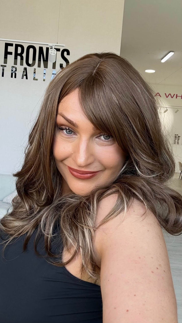 Woman smiling in a warm brunette wig with honey blonde highlights, worn with a middle part and a long fringe parted to the side.