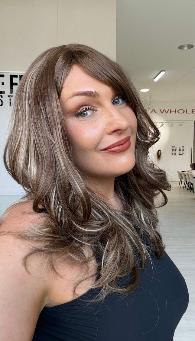 Smiling woman wearing Suki, a warm brunette wig with ash and honey blonde highlights throughout and a long fringe parted to the side.