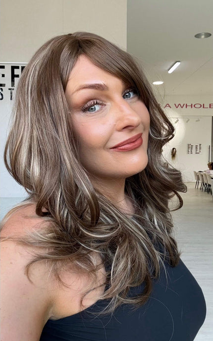 Smiling woman wearing Suki, a warm brunette wig with ash and honey blonde highlights throughout and a long fringe parted to the side.