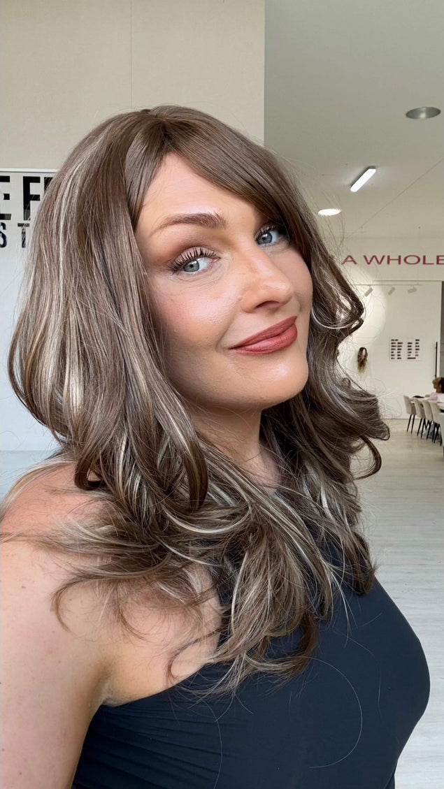 Smiling woman wearing Suki, a warm brunette wig with ash and honey blonde highlights throughout and a long fringe parted to the side.
