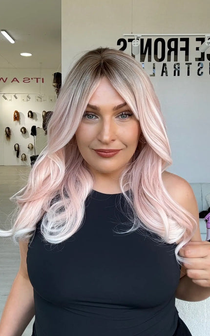 a soft pastel pink mid length wig with  a middle part and darker shadow root, hair comes to mid chest length and is perfect for daily wear with a glueless wig cap, and heat safe/ styleable hair.