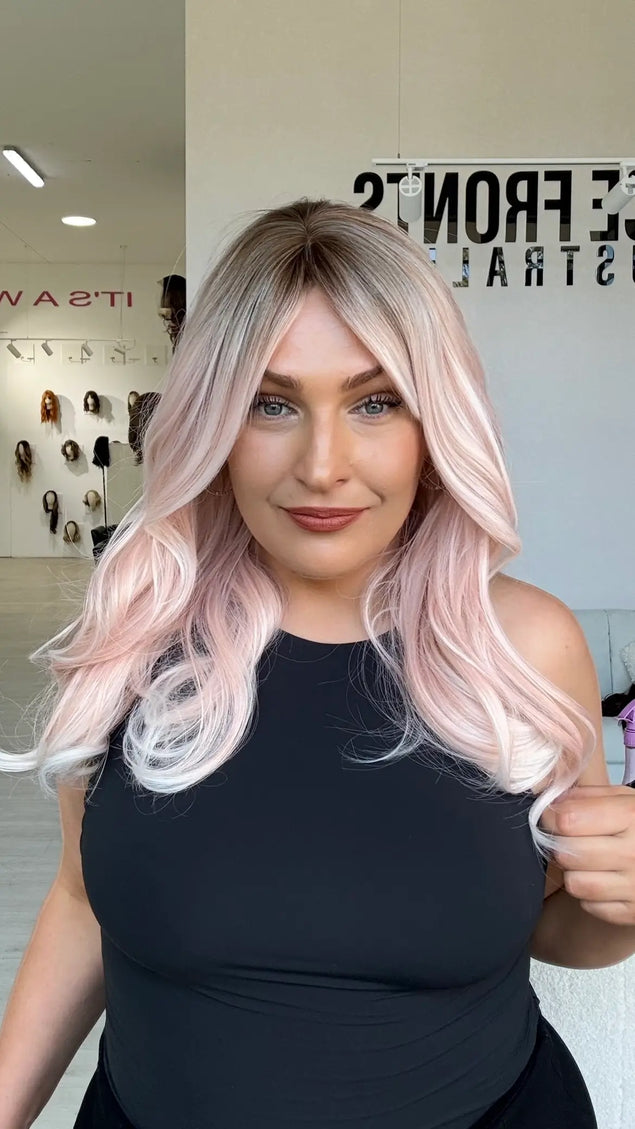 a soft pastel pink mid length wig with  a middle part and darker shadow root, hair comes to mid chest length and is perfect for daily wear with a glueless wig cap, and heat safe/ styleable hair.