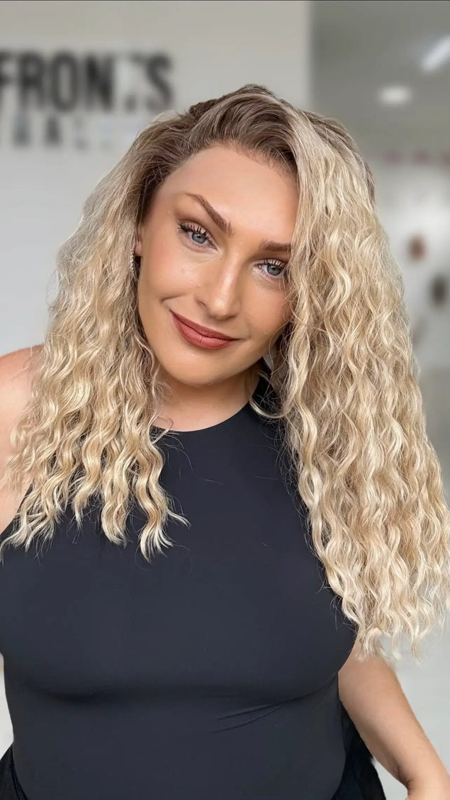 a side part on the curly honey blonde hair with soft brunette shadow root, lace front wig with natural looking hairline, designed for easy wear.