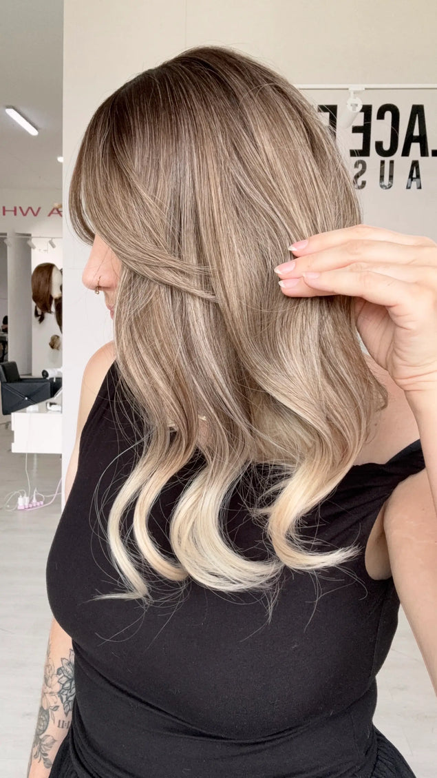 side view showing ash blonde ombre, darker root to ash mids and platinum ends. has a slight curl on ends, middle part and long curtain bang fringe.