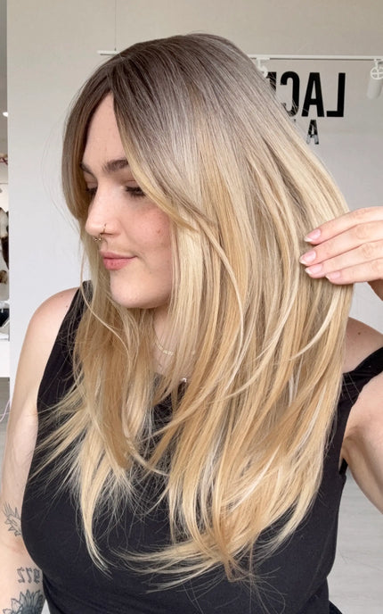 side view holding the layered vanilla blonde hair with a soft brunette root. layers throughout for movement, a middle part and long curtain bangs parted to the side.
