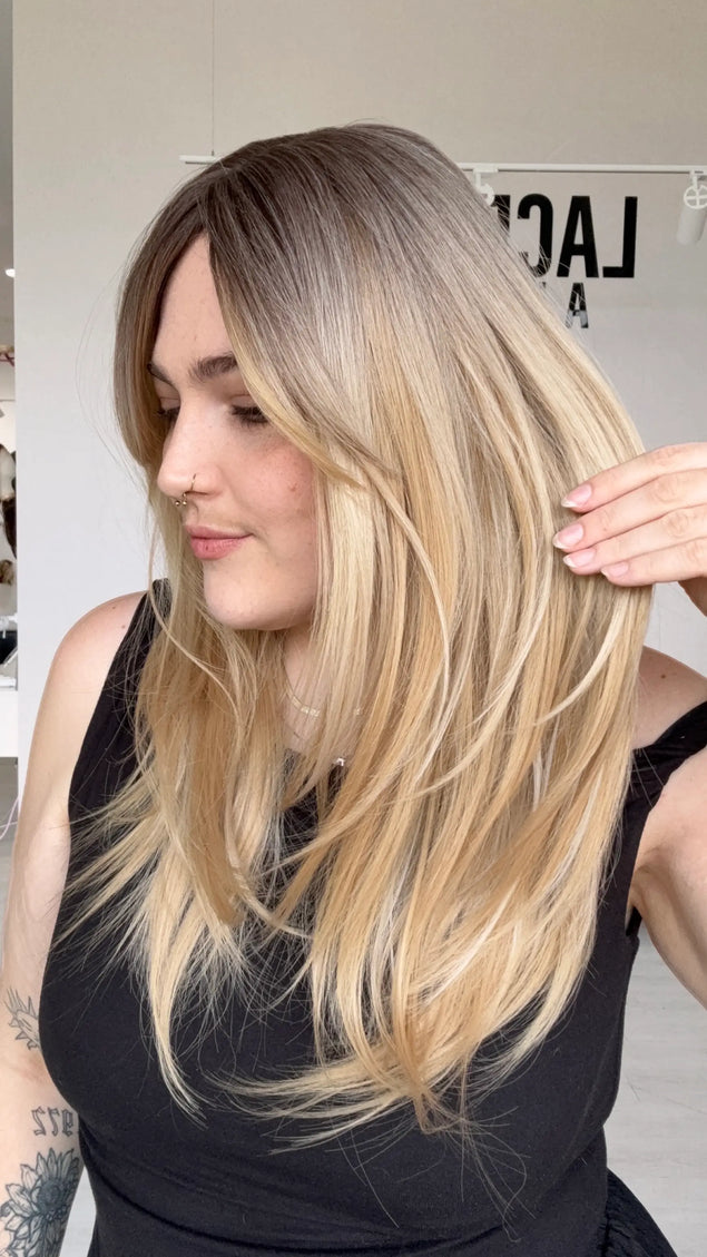 side view holding the layered vanilla blonde hair with a soft brunette root. layers throughout for movement, a middle part and long curtain bangs parted to the side.