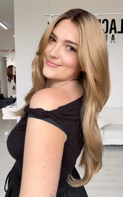 side view showing long length layered honey blonde hair, featuring a dark shadow root to dark honey blonde hair, this wig has a set middle part.