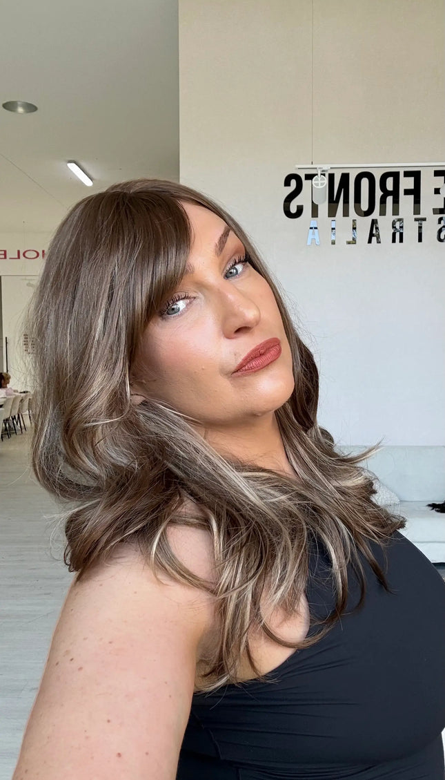Warm brunette hair with ash and honey blonde highlights, light layers throughout, a middle part and a long fringe parted to the side.