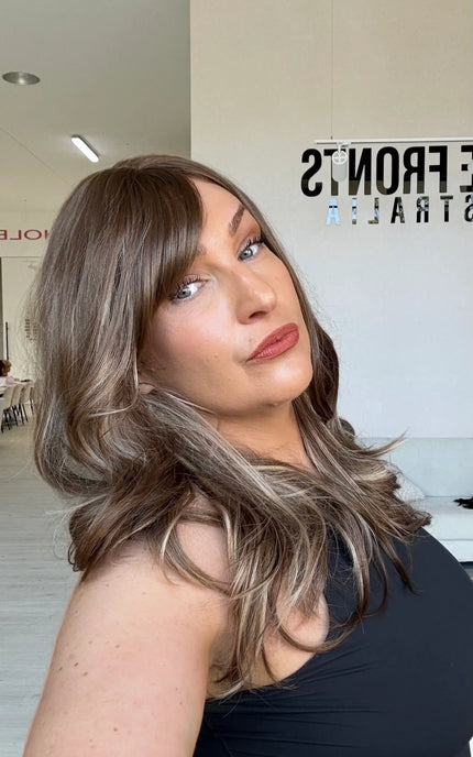 Warm brunette hair with ash and honey blonde highlights, light layers throughout, a middle part and a long fringe parted to the side.