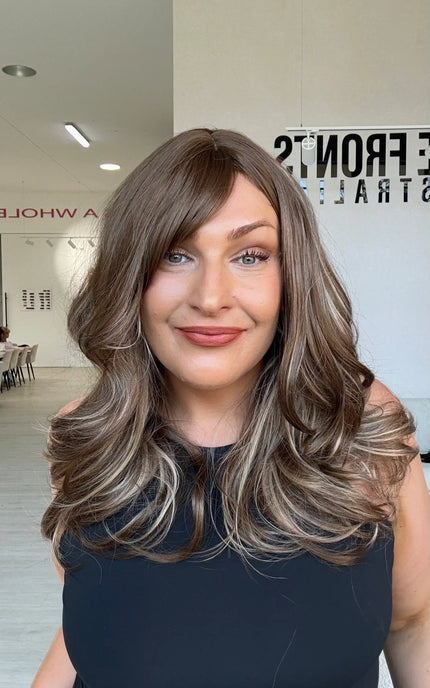 Woman in a black singlet wearing Suki, a warm brunette wig with ash and honey blonde highlights underneath, a middle part and a long fringe swept to the side.
