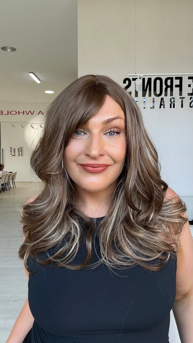 Woman in a black singlet wearing Suki, a warm brunette wig with ash and honey blonde highlights underneath, a middle part and a long fringe swept to the side.